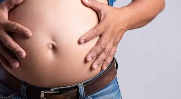 Un homme, je suis mince avec un gros ventre, tient son abdomen proéminent au-dessus de son jean et de sa ceinture.