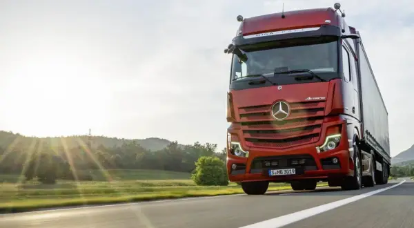 Un camion rouge vif roule sur une route ensoleillée, le soleil couchant illuminant le paysage. Ce camion sans rétroviseur semble prêt pour une longue route.