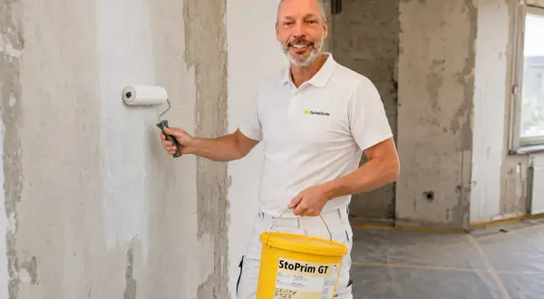 Maler grundiert mit einer Rolle die Wand. Er hält einen Eimer mit "StoPrim GT" in der Hand.