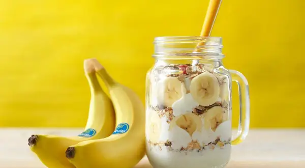 Un delizioso barattolo con yogurt, granola e fette di banana, perfetto per una banana a colazione. Accanto, due banane fresche su sfondo giallo.