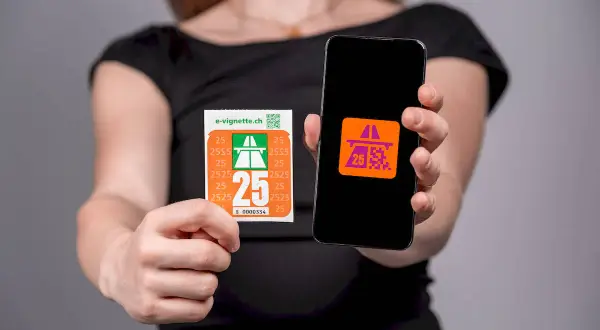 Ręce trzymają naklejkę z napisem "e-vignette.ch" i numerem "25", obok telefonu z aplikacją symbolizującą winietę szwajcaria.