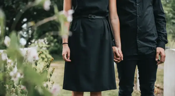 Ein Paar in schwarzer Kleidung, die Frau in einem schlichten Kleid, hält Händchen. Passende **Beerdigung Kleidung Frau Sommer** für einen traurigen Anlass.