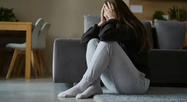 Kobieta siedzi na podłodze, zakrywając twarz dłońmi. Jej postawa sugeruje przygnębienie i objawy stresu długotrwałego.