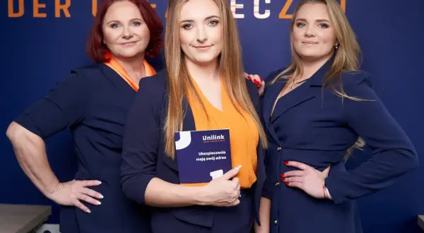Trzy kobiety w granatowych marynarkach i pomarańczowych bluzkach pozują na tle niebieskiej ściany z napisem.
