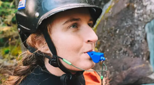 Persona con casco y traje naranja sopla un silbato de emergencia azul, listo para cualquier aventura acuática.