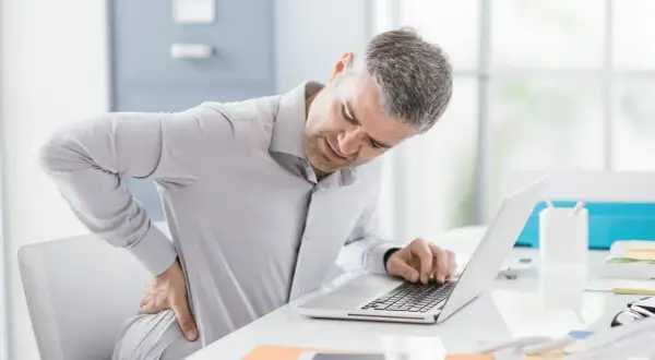 Mężczyzna z bólem pleców przy biurku, pracujący na laptopie. To może być objaw choroby zawodowej.