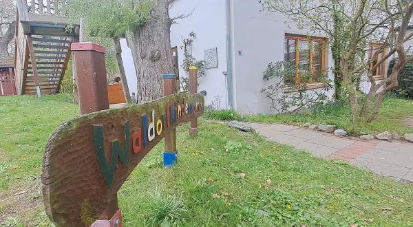 Holzschild "Waldorfkindergarten" vor einem Gebäude mit Spielturm. Mögliche Nachteile: wenig Struktur, viel Freiheit.