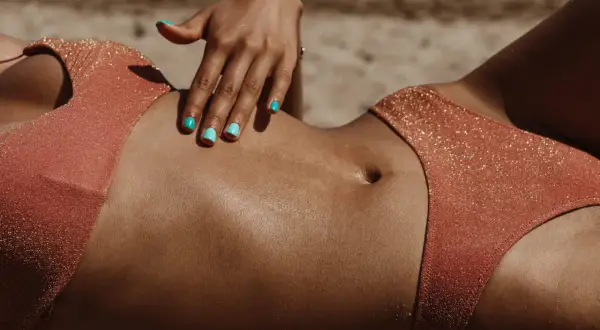 Main d'une femme aux ongles turquoise caressant son ventre bronzé. Une question se pose : combien de temps dure un bronzage ?