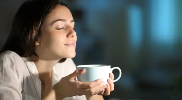 Kobieta z zamkniętymi oczami wdycha aromat napoju. Może to być kawa lub napar z ziół na pobudzenie.