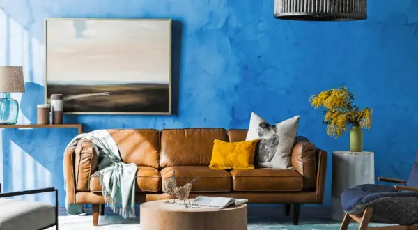 Ein Wohnzimmer mit leuchtend blauer Wand. Ein braunes Ledersofa, ein gelbes Kissen und ein Bild mit Landschaftsmotiv. Welche Farbe passt gut zu Blau? Hier: Gelb und Braun.