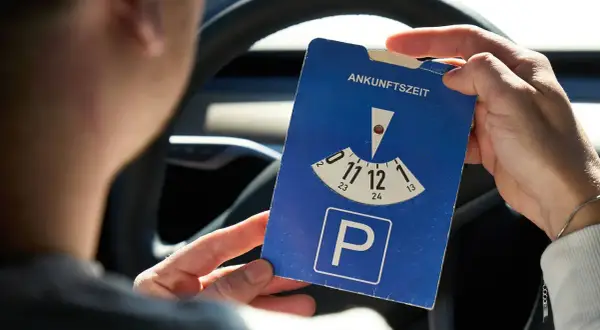 Person hält eine Parkscheibe mit der Aufschrift "Ankunftszeit" und dem Parkplatzsymbol.