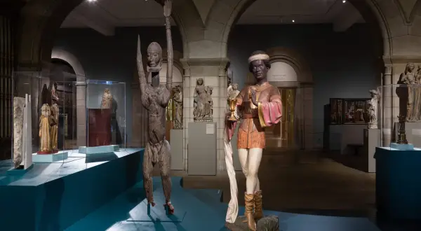 Two striking sculptures, one a wooden figure with raised arms, the other a painted figure holding a chalice, are displayed in a museum gallery, showcasing medieval African art.