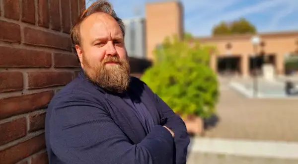 Nicolas Ballario, con barba folta e sguardo intenso, appoggiato a un muro di mattoni rossi, con un blazer blu.