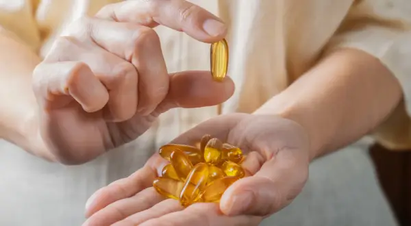 Dłoń trzyma kapsułkę z omega-3, zarejestrowaną jako lek. W drugiej dłoni garść takich tabletek.