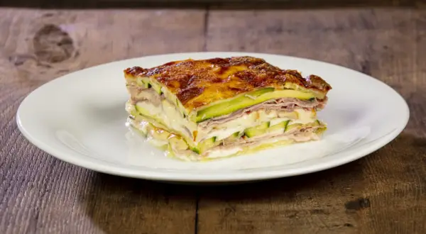 Una fetta di parmigiana di zucchine bianca alla napoletana, stratificata con prosciutto e formaggio fuso, su un piatto bianco.