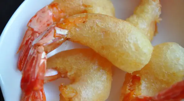 Plusieurs beignets de crevette dorés et croustillants, avec des queues de crevettes roses qui dépassent, servis dans une assiette blanche.