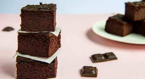 Ciemne, kwadratowe brownie ułożone w stos, obok kawałki czekolady na różowym tle.