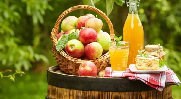 Frischer, selbstgemachter Apfelsaft in einer Flasche und einem Glas, daneben ein Korb voller Äpfel und ein Stück Apfelkuchen. Haltbarkeit von selbstgemachtem Apfelsaft beachten.
