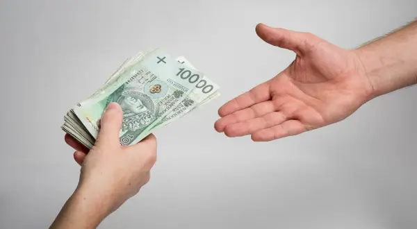 Dłoń podaje plik banknotów 100 zł, druga dłoń wyciągnięta do odbioru. Ilustracja tematu odsetki ustawowe za opóźnienie w transakcjach handlowych.