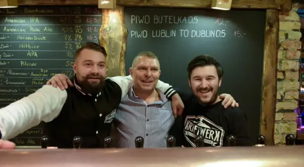 Trzech uśmiechniętych mężczyzn za **tap barem** w pubie. Na tablicy za nimi widnieje menu piwne.