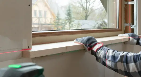 Precyzyjny montaż parapetu drewnianego z użyciem poziomicy laserowej. Ręce w rękawicach przytrzymują deskę, a czerwone linie lasera wyznaczają idealny poziom.