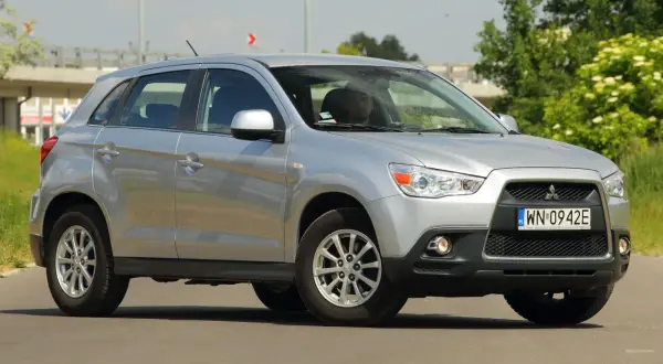 Srebrny Mitsubishi ASX 1.6 benzyna z kierowcą za kierownicą, zaparkowany na drodze z zielenią w tle.