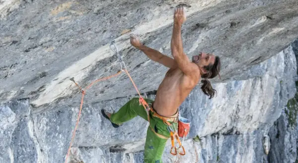 Alexander Huber klettert an einer steilen Felswand, sein Körper angespannt, die Hände greifen nach kleinen Vorsprüngen.