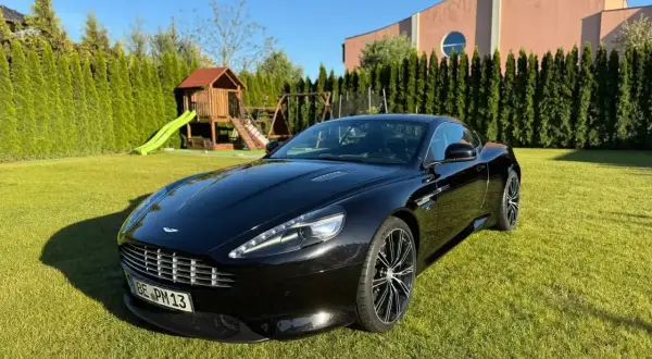 Czarny Aston Martin DBS lśni na trawniku przed domem. W tle plac zabaw i wysokie tuje.