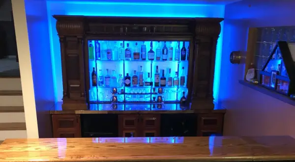 A stunning home bar setup, perfect for entertaining. This image shows how to build a bar with a wooden countertop, a backlit liquor cabinet filled with bottles, and vibrant blue ambient lighting.