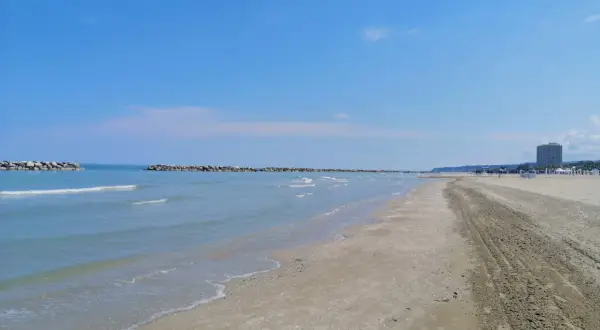 Spiaggia del lido di Fermo con mare calmo, frangiflutti in pietra e un palazzo sullo sfondo.