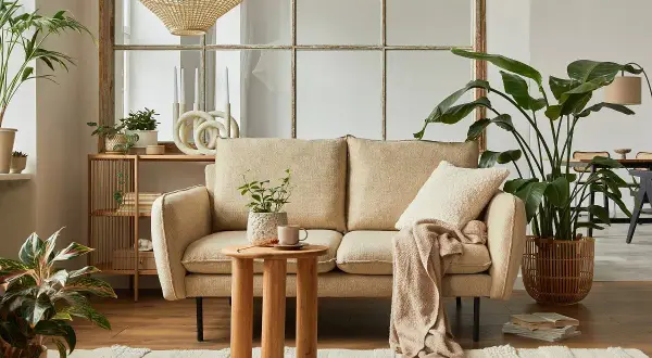 Przytulny salon w stylu cottage: beżowa kanapa, drewniany stolik, dużo roślin i pleciony klosz lampy tworzą ciepłą atmosferę.