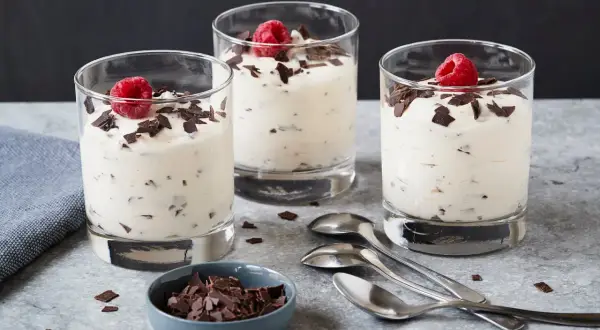 Drei Gläser mit cremigem Dessert, garniert mit Himbeeren und Schokoladenstückchen. Ein Schälchen mit Schokostückchen und Löffel daneben. Ein leckeres Herrencreme Rezept zum Nachmachen.