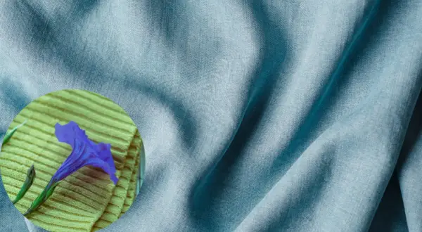 A blue tencel fabric drapes softly, hinting at its smooth, natural feel. A purple flower rests on a textured yellow surface.