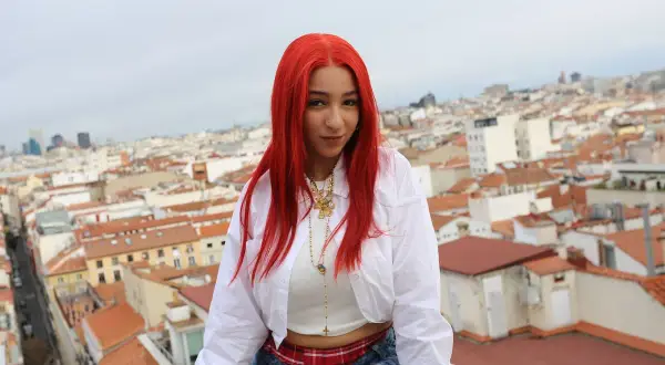 Joven con cabello rojo vibrante, como una rosa encendida, posa sobre tejados urbanos.