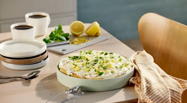 En krämig tiramisu limoncello, garnerad med citronzest och persilja, står på ett bord med kaffe och citroner.