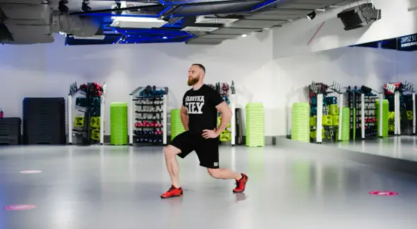 Mężczyzna wykonuje wykroki, ćwicząc odwodziciele uda. W tle widać sprzęt do ćwiczeń w siłowni.