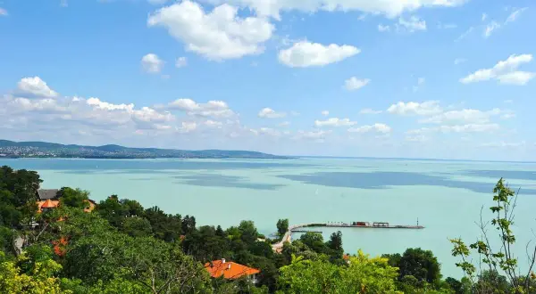 Widok na turkusowe jezioro Balaton z zielonymi wzgórzami i domami. Na wodzie widać molo i żaglówki.