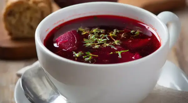 Aromaticzny barszcz czerwony domowy, z kawałkami buraka i ziołami, podany w białej filiżance z łyżeczką.