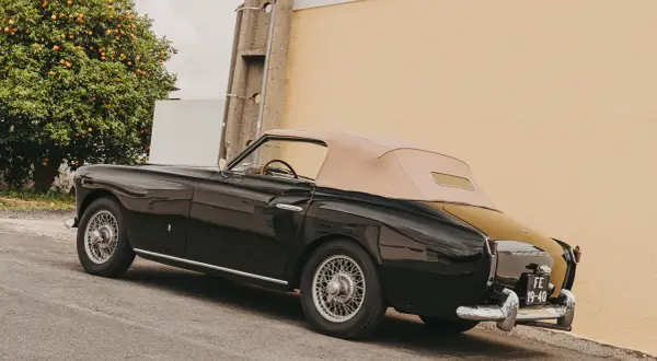 Svart, klassisk brittisk sportbil med beige sufflett parkerad vid en gul vägg, bredvid ett apelsinträd.