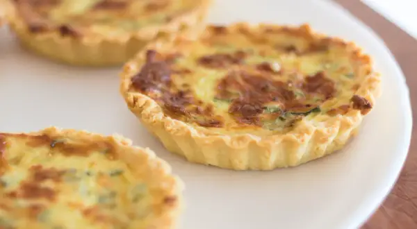 Trois petites quiches sans œuf dorées sur une assiette blanche. La pâte est croustillante et la garniture semble crémeuse.