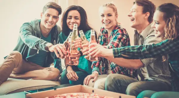 Grupa przyjaciół wznosi toast butelkami piwa, śmiejąc się i jedząc pizzę. Nawet trudne pytania nie psują im nastroju.
