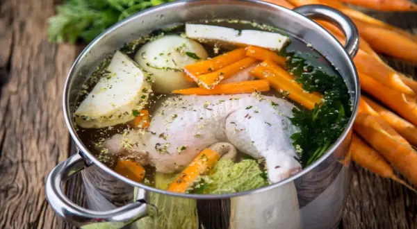 Pentola con brodo di pollo ricetta della nonna: pollo, carote, sedano, cipolla e prezzemolo in cottura su un tavolo di legno.