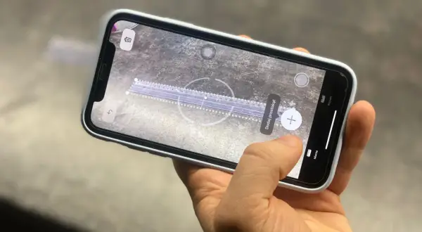 Dłoń trzyma telefon z włączoną aplikacją miarka. Na ekranie widoczna jest wirtualna linijka i przycisk "Dodaj prostokąt".