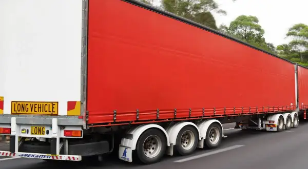 Un long camion 60 tonnes, avec une remorque rouge vif, roule sur une route bordée d'arbres.