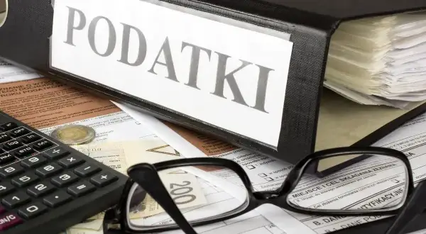 Segregator z napisem "PODATKI", okulary, kalkulator i pieniądze na dokumentach. Czas na rozliczenie PIT-2.
