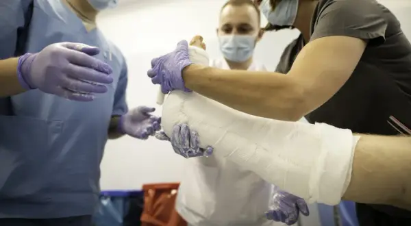 Trzy osoby w maseczkach i rękawiczkach zakładają gips na nogę pacjenta.
