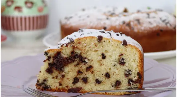 Una fetta di torta soffice con tante gocce di cioccolato, cosparsa di zucchero a velo, su un piatto lilla con una forchetta.