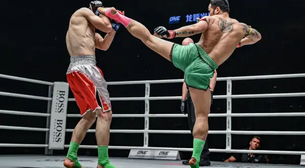 A Muay Thai fighter in green shorts lands a powerful kick to his opponent's head. The opponent, in red shorts, braces for impact, his foot nearly touching the canvas.