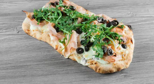 Pinsa co to? Podłużna, chrupiąca baza z szynką, mozzarellą, rukolą i oliwkami. Pyszna alternatywa dla pizzy.