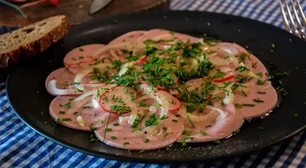 Saure Wurst mit Zwiebeln, Radieschen und Schnittlauch auf schwarzem Teller, dazu Brot.